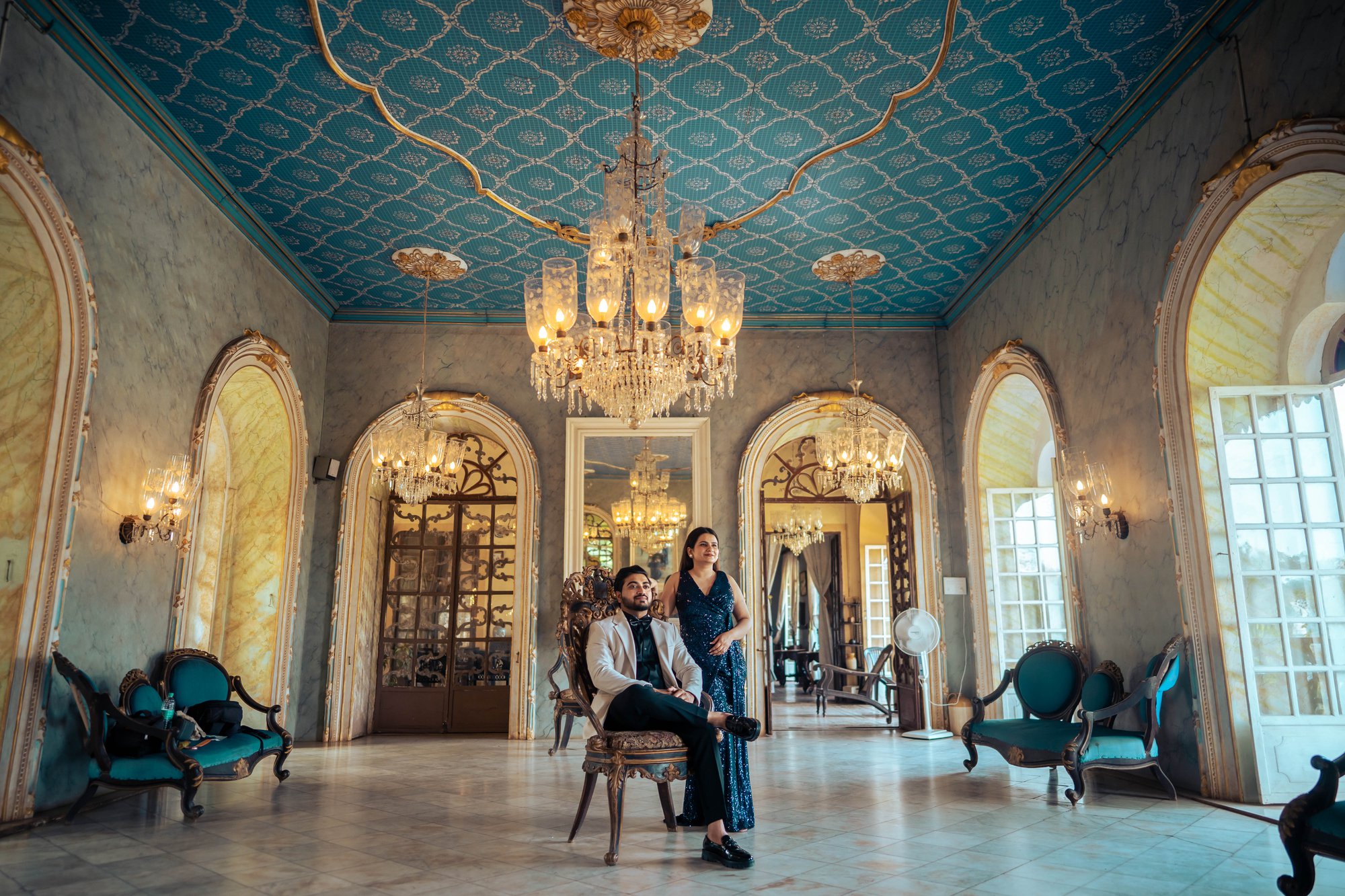 Regal pre-wedding shot—groom sitting gracefully while bride stands beside him in a magnificent hall.